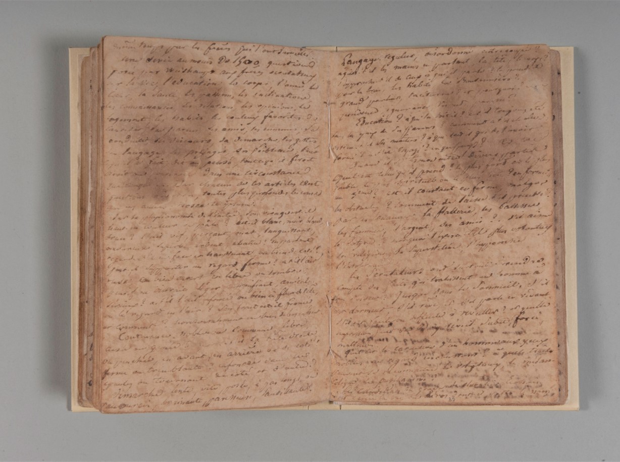 This image is a photograph. The photograph features Aimé-Adrien Taunay's notebook open on a gray surface. There are handwritten inscriptions in iron gall ink and moisture stains on the leaves.